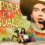Johanny sits under a mural of a colorful woman with the words "Poder E Igualdad"