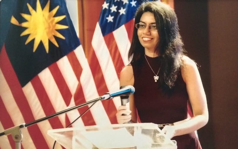 Hervinder presenting at the Ministry of Higher Education Malaysia during the Fulbright Conference standing behind a microphone with a the Malaysian and US Flags in the background